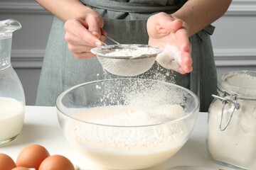 Making batter (liquid dough). Woman sieving flour into bowl at white table indoors, closeup