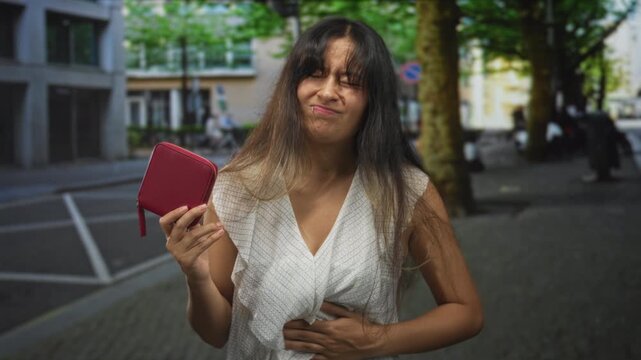 Young latina brunette woman clutching stomach and holding a red wallet on a city street with trees and buildings; financial stress pain.