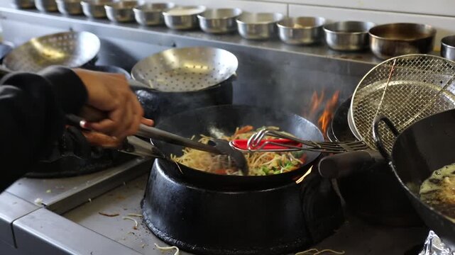 Chef stir frying Asian noodles with vegetables in hot wok at restaurant kitchen, with steam and flame.