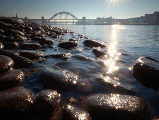 Riverside stones low angle view, bright sunburst on rippling water, arch bridge background, tranquil city scene. Clear sky, urban waterfront.