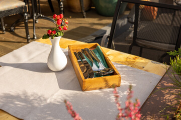 Cutlery set in wooden box and white vase with red berries on outdoor restaurant table.