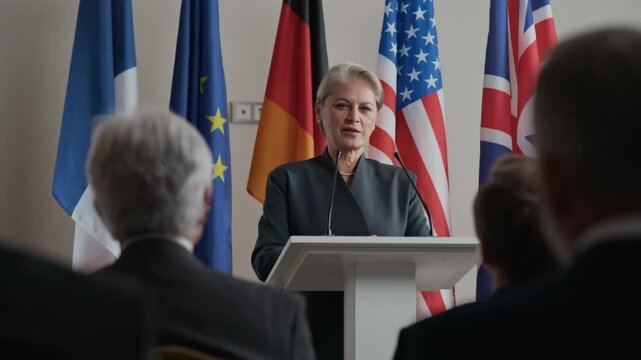 Medium shot of confident woman delivering speech at podium with multiple national flags behind her symbolizing international politics and global dialogue