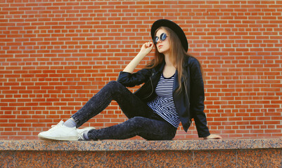 Stylish young woman model wearing black round hat, leather jacket in rock style on city street