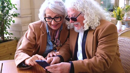 Happy senior couple sitting together at outdoor cafe using smartphone and looking at social media. Elderly people enjoying technology, connection, communication, digital lifestyle, love and leisure - Powered by Adobe