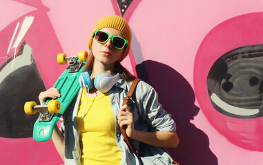 Stylish modern young teenager girl with skateboard in glasses, listening to music, graffiti wall