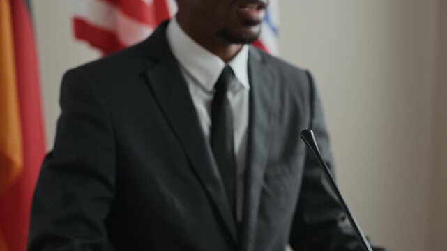 Tilt up of confident Black male politician in suit speaking at podium with microphones, addressing press and audience during formal political or international event