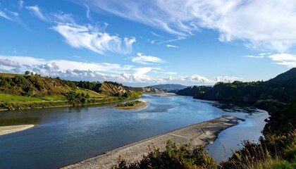 Expansive scenic view of a wide river with a sandy bank, surrounded by rolling green hills under a partly cloudy sky