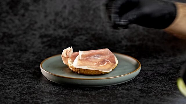 Male hand in glove assembling delicious bagel sandwich on teal plate. Layers of ham, avocado, pickles, and egg topped with sesame and spices. Dark background, 
food preparation concept.