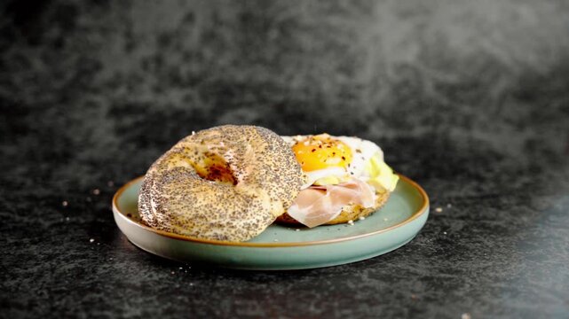 Delicious breakfast bagel sandwich with fried egg, prosciutto, and avocado, topped with sesame and poppy seeds. Served on matte teal plate, side view, dark background, gourmet food concept.
