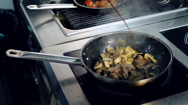 Dark sauce being added to saut&eacute;ed mushrooms and meat in frying pan. Steam rising, second pan with tomatoes in background.