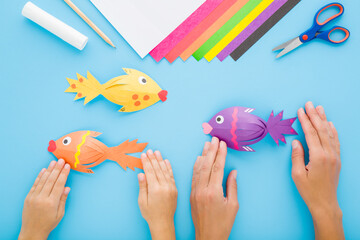 Mother and little child hands creating fish shapes. Scissors, glue stick, pencil and colorful application paper. Blue table background. Pastel color. Flat lay. Point of view shot. Top down view.