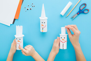 Mother and child hands together making and decorating snowman shapes from white paper on blue table background. Pastel color. Flat lay. Point of view shot. Creating winter elements. Top down view.