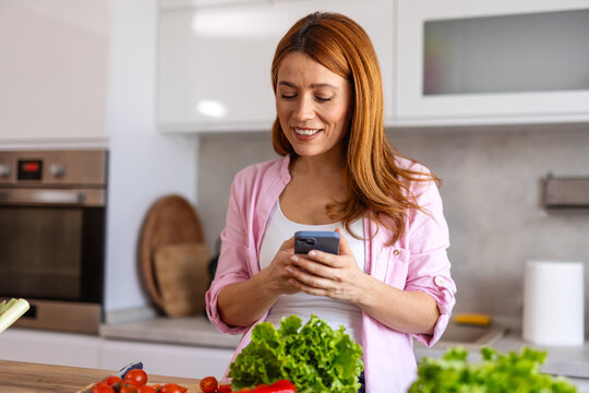 A woman in a modern kitchen prepares dinner while taking food photo, capturing a fun and joyful moment. She is multitasking, cooking a delicious homemade meal while enjoying the process. - Powered by Adobe