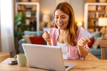 A happy woman enjoys online shopping at home, holding a credit card while browsing on her laptop in...