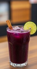A glass of purple drink with ice cinnamon sticks and a lime slice on a wooden surface indoors