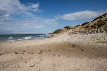 A scenic coastal landscape featuring a sandy beach bordered by gentle waves and a deep blue sea. A grassy, sunlit cliff along the shoreline under a partly cloudy sky, peaceful and natural seaside
