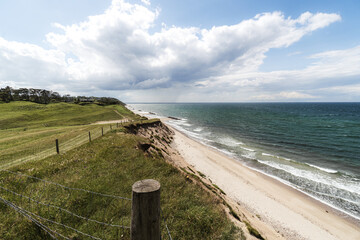 Windswept coastal scene, a grassy cliff overlooking a sandy shoreline and deep blue waves. A wooden fence lines the edge of the bluff, dramatic clouds across the bright sky,  serene seaside atmosphere