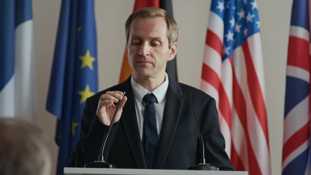 Dolly-in of confident male official addressing journalists during international political event, standing at podium with microphone, surrounded by flags of multiple nations