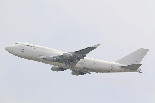 An unmarked, all-white large cargo jet airplane climbing after takeoff against a plain overcast sky. - Powered by Adobe