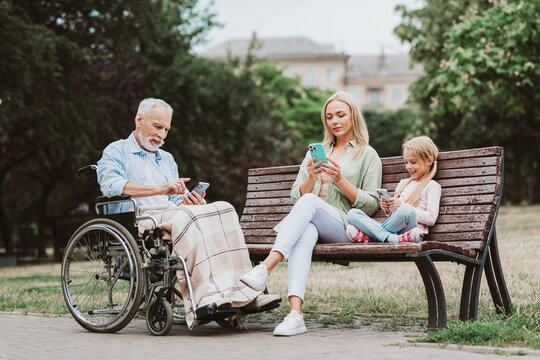 Family scene at the park with three generations using phones on a bench as grandpa in a wheelchair shares a moment with his daughter and granddaughter