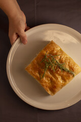 A hand is presenting a square baked dish that is beautifully garnished with a fragrant herb on a plate