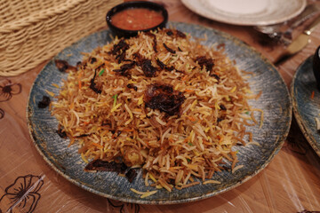 A detailed, close-up shot of a large, rustic blue ceramic plate piled high with aromatic Middle Eastern or South Asian rice, such as Biryani or Mandi. The long-grain basmati rice is topped with crispy