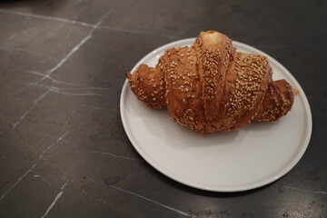 A close-up, high-angle view of a golden-brown, flaky croissant generously topped with toasted sesame seeds. The pastry is served on a simple white ceramic plate, which sits on a dark, elegant, veined 