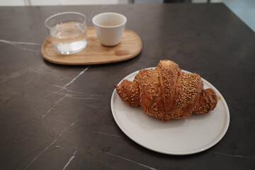 A golden-brown, flaky croissant covered in toasted sesame seeds is served on a simple white plate. In the blurred background, a small wooden tray holds a glass of water and a white coffee cup.