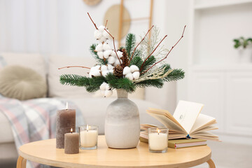 Winter composition with fir tree branches, cotton flowers in vase, books and burning candles on wooden table indoors, closeup
