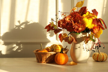 Autumn atmosphere. Vase with beautiful flowers, pumpkins and berries on wooden table near light wall, space for text