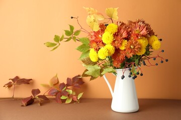 Autumn atmosphere. Vase with beautiful flowers, berries and leaves on brown table against orange background
