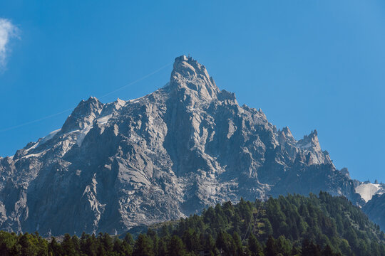 Aiguille du Midi, a prominent mountain peak in the Mont Blanc massif of the French Alps. Majestic rocky mountain peak with cable car and forested slopes under a clear blue sky.