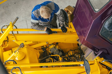 A mechanic in work gloves examines the engine of heavy machinery in a workshop. The setting is industrial, showcasing maintenance and repair work.