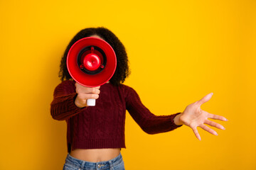 Obraz premium Young mixed race woman with curly hair holds a red megaphone and extends a hand against a bright yellow background promoting fun