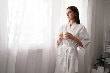  beautiful young woman in a white bathrobe wakes up in the morning, standing by the window drinking coffee, looking outside at the curtains in the bedroom in the morning.

