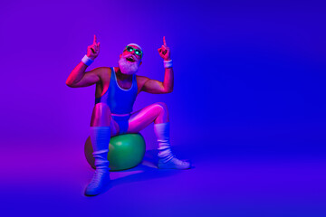 Fototapeta premium Retro fitness man in bright blue jumpsuit sits on a green exercise ball under neon purple and blue lights posing with raised fingers