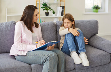 A child psychologist conducts an assessment while taking notes on a clipboard as the young girl sits tense on the sofa in therapy. Therapist asks gentle questions and listens closely.