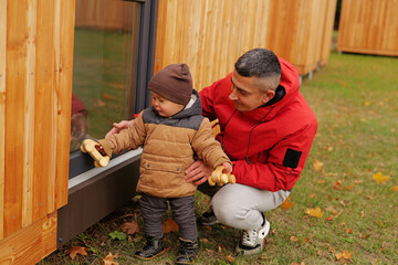 A father shares joyful moments with his young son outdoors, playing with toys and exploring their grassy lawn near their wooden home on a bright day