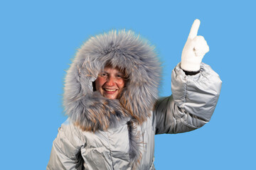 A woman wearing a winter coat with a fur-lined hood smiles and points upwards with her gloved hand. There is copy space above her finger.