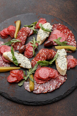 Various sliced dry - cured Italian sausages. Cold cuts with olives, bruschetta, marinated cucumber, arugula and basil leaves on black slate plate. Close up, overhead view