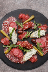 Various sliced dry - cured Italian sausages. Cold cuts with olives, bruschetta, marinated cucumber, arugula and basil leaves on black slate plate. Close up, overhead view