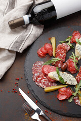 Close up view of antipasto meat platter. Cold cuts with olives, croutons, marinated cucumber, arugula and basil leaves on black slate plate with fork, knife and bottle of wine