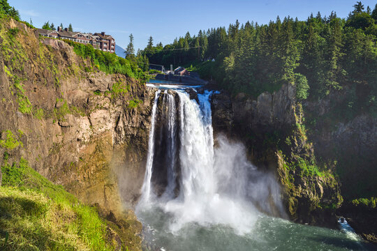 A scenic view of the powerful Snoqualmie Falls cascading over cliffs, with the historic Salish Lodge & Spa visible on the cliffside above, all set against a clear blue sky.