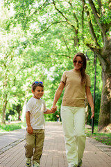 Obraz premium Front view of mother walking down the street with little son with sunglasses on sunny day in public green park.