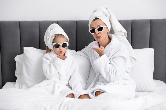 Mother and daughter in bathrobes with towels wrapped around hair and sunglasses blowing air kisses on bed at home