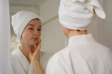 beautiful young woman wearing a bathrobe and using a towel to cover her hair after taking a shower looks at her face in the mirror and applies facial serum in her bedroom at home.