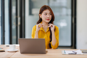 Smiling asian female accountant looking away while analyzing financial report over laptop at table in office