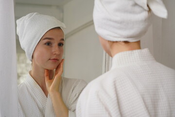 Young woman wearing a bathrobe and a towel covering her hair, looking in the mirror in the morning, getting dressed and applying cream in her bedroom at home.
