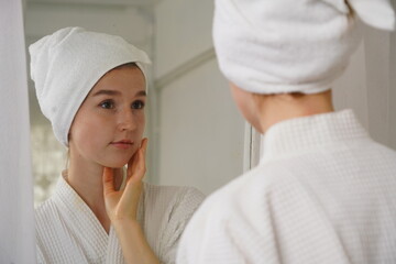 Young woman wearing a bathrobe and a towel covering her hair, looking in the mirror in the morning, getting dressed and applying cream in her bedroom at home.

