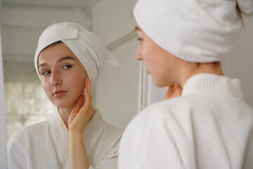Beautiful young woman wearing a bathrobe and using a towel to cover her hair after taking a shower, looks in the mirror and sees a happy smiling face with fresh skin in the dressing room at home.